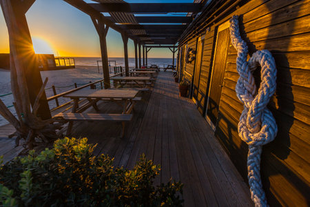 A seaside building with a wooden deck, tables, and a rope knot faces the ocean at sunset.の写真素材