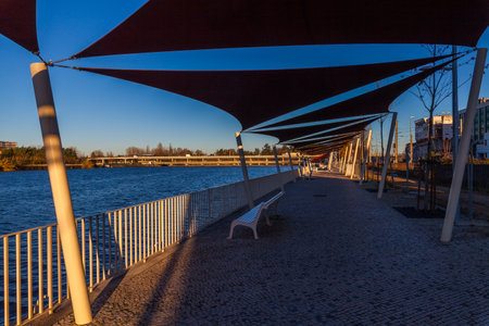 A paved walkway with shade sails runs alongside a river, offering a scenic view of the water and distant hillsの写真素材