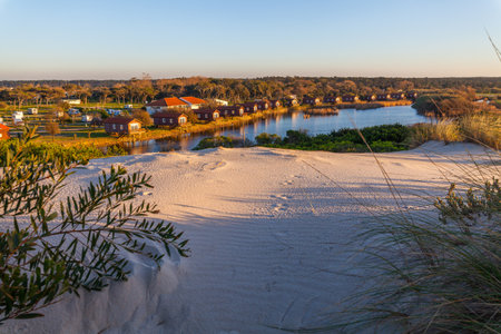Sand dunes and vegetation overlook a waterway lined with buildings and trees under a clear sky.の写真素材
