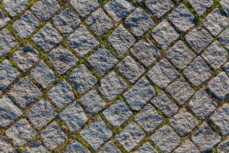 A textured cobblestone road surface, with tufts of grass growing between the stones, is illuminated by warm sunlight casting long shadows, creating a rustic and slightly weathered sceneの写真素材