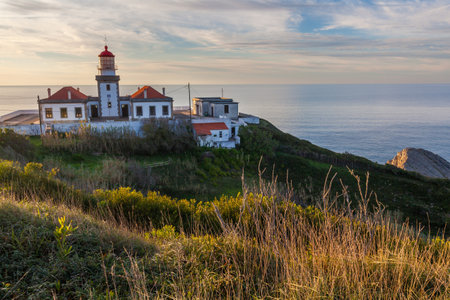 A white lighthouse with a red roof stands prominently on a coastal promontory, with a paved pathway leading up to it under a blue sky streaked with wispy cloudsの写真素材