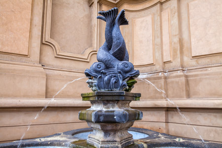 A decorative fountain featuring two intertwined fish or dolphin figures atop a pedestal, with water flowing from their mouths into a basin below, set against an architectural backdrop.の写真素材