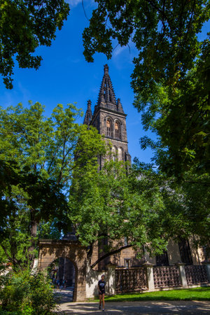 A gothic church tower with spires rises above a stone wall and arched gate, framed by lush green trees and a clear blue sky, with a person walking in the foreground.の写真素材