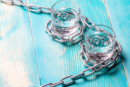 Two glasses of vodka on a wooden painted table are connected by a chain of metal, glass and metalの写真素材
