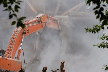 through the trees on the foreground is an excavator bucket to break the building, dismantling of damaged buildings, dust and stones. destruction of buildingsの写真素材