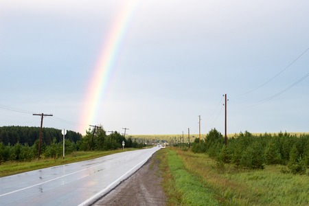 countryside road wet with rain and part of the rainbow over the road, cloudy skyの写真素材