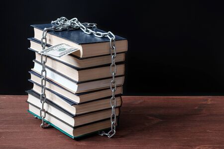 a rickety stack of books standing on a wooden surface is tied with a metal chain sticking a dollar and the top of the bookの写真素材