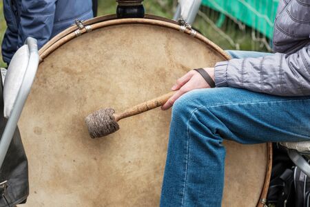 big drum and drummer sitting next to him with a stick outside. street musician with street clothes