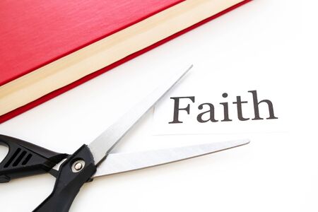 scissors open for cutting and a strip of paper with the word faith on a white background next to a red bookの写真素材