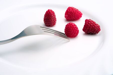 unfocused metal fork and close-up of four ripe raspberries on a white ceramic surface
