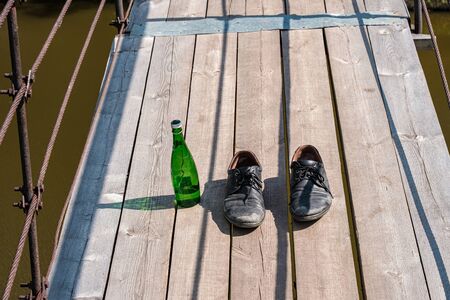 dirty old shoes on a wooden suspension bridge over the river nearby is a green glass bottle. drank and jumped out to swimの写真素材