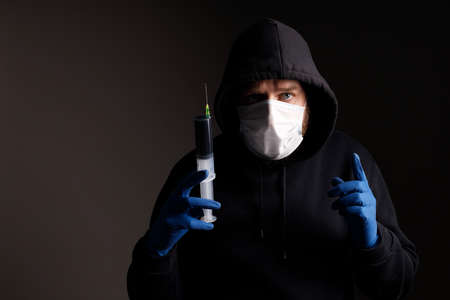 a white man with a beard, wearing a medical mask and rubber gloves, is holding a large syringe with a black liquid on a dark background. medicine or drug in hand warning of dangerの写真素材