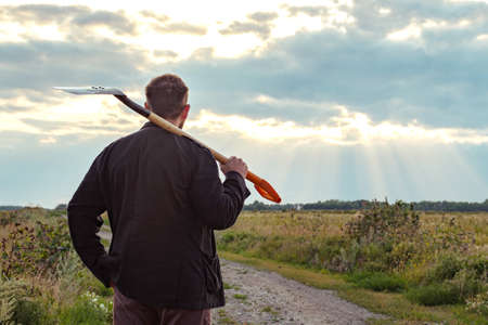 a man with a shovel on his shoulder stands with his back looking into the distance on a field road...