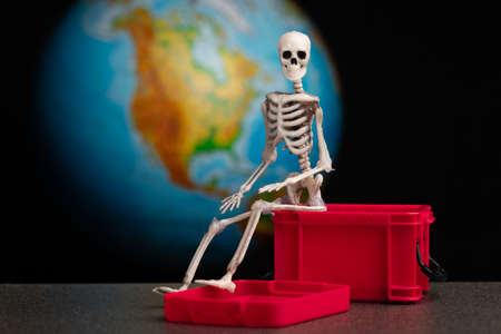 the skeleton sits on a red plastic box that is open, background planet earth map of north america. the concept of high mortality and global problemsの写真素材