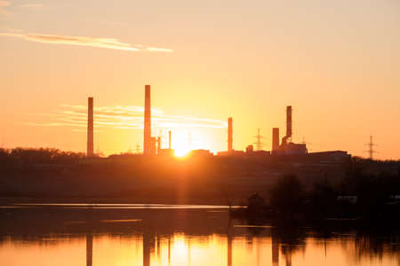 rays of the sun through the factory chimneys during sunset on the shore of the reservoir. pipes are reflected in the waterの写真素材