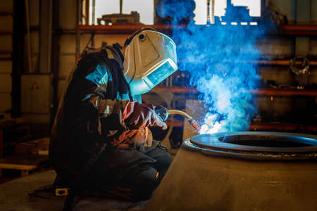 a welder at work welds a round part illuminated by a mask from the work of the welding machine. metal welding in the factoryの写真素材