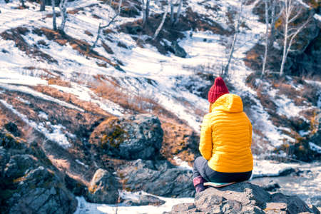 girl in a yellow jacket and a red knitted hat in nature sits on a stone. rest in the highlands in spring. reflections on life in nature away from the city. place of powerの写真素材