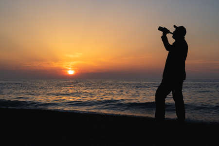 a man in a cap drinks wine from a bottle during a sunset at the sea. silhouette of a drinking man on...
