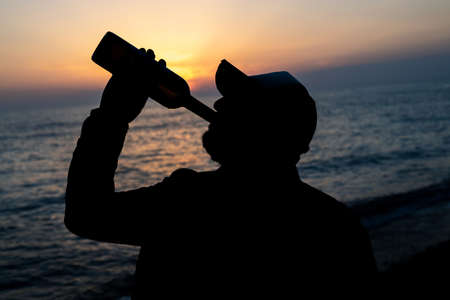 blurred silhouette of a man by the sea drinking wine straight from a bottle against the backdrop of the sunset sky. vacation at seaの写真素材