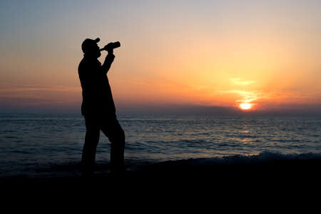 silhouette of a lonely man on the background of the sea at sunset time. man drinks wine from a bottle. loneliness and a walk to the seaの写真素材