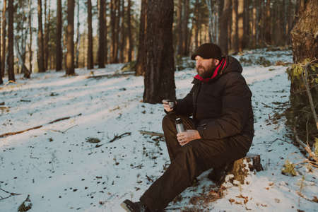 an adult bearded man sits in the forest with a mug of tea and a flask . looks thoughtfully in front of himの写真素材