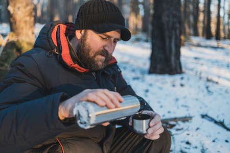 a bearded man sits in a winter forest and pours himself tea from a flask . nature travelerの写真素材