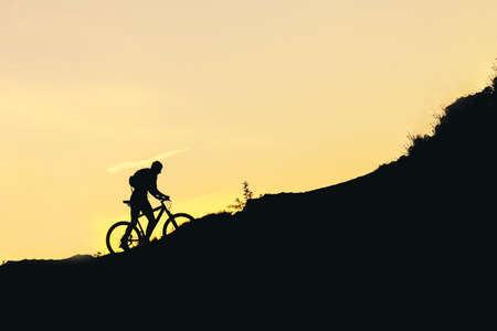 silhouette of a man on a bicycle going uphill during sunset. summer evening overcoming difficultiesの写真素材