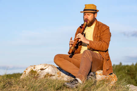 adult bearded man closing his eyes plays the flute while sitting in nature. outdoor musicianの写真素材