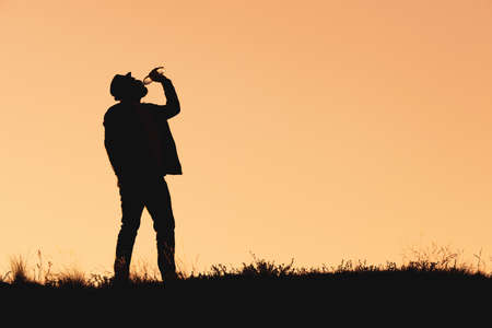 silhouette of a man in a hat who drinks water from a bottle. thirst quencher, outdoor orange backgroundの写真素材