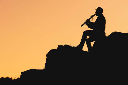 silhouette of a man in a hat sitting on a mountain and playing the flute. musician outdoors playing in natureの写真素材