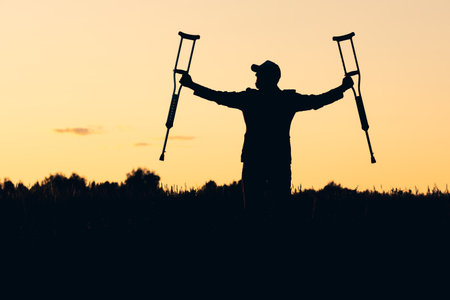 silhouette of a man in a cap during the dawn on the field with crutches in his hands. recovery and recovery, free movementの写真素材