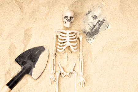 shovel in the sand skeleton and one hundred dollars. Franklin portrait peering out from under the sandの写真素材