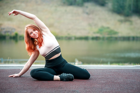 doing sports outdoors on a summer day against the backdrop of nature, a red-haired girl in sportswear sits and pulls her handの写真素材