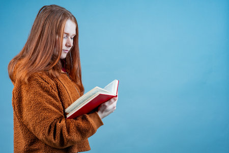 a young woman stands in the studio on a blue background and reads a red book. careful reading of...