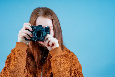 a young girl with a camera on a blue background in a brown jacket. photographer with loose red hairの写真素材