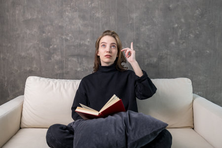 A young woman is sitting on a modern white sofa reading a book and holding up her finger. the idea...