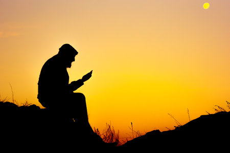 the silhouette of a man sitting on the ground and reading a book. a man reading against the background of a sunsetの写真素材