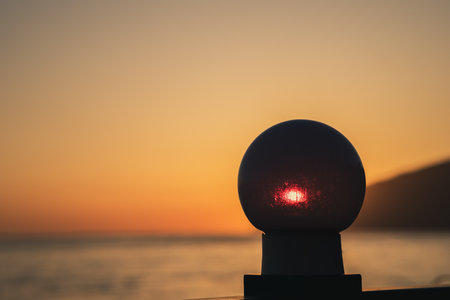 A glowing ball sits on a post by the ocean. The sun is setting, casting a warm glow over the scene. The ball's light seems to be reflecting off the water, creating a serene and peaceful atmosphereの写真素材