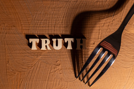 a metal fork on a brown background and a word made of wooden letters - the truth, the fork casts a crooked shadow on the word truth.の写真素材