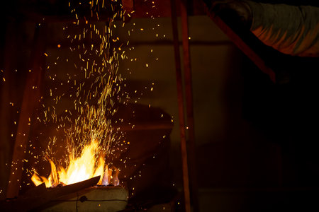A man is standing over a fire, with sparks flying up into the air. The scene is dark and moody, with...