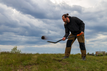 A man is digging in the grass with a shovel. The sky is cloudy and the man is wearing a black jacketの写真素材