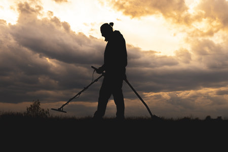 The man searches for metal objects in the ground with a metal detector, and the man has a shovel in his other hand. Sky with clouds before rainの写真素材