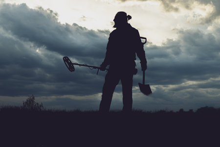 A man stands in the woods with a metal detector and a shovel. The sky is covered with clouds, and the silhouette of a man with a beard is visible against the background of the sky.の写真素材