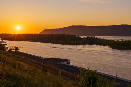 The sun is setting over a river, casting a warm glow over the water. The sky is a mix of orange and...