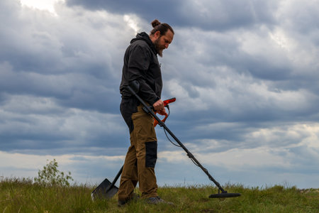 A man with a beard searches for metal objects against a cloudy sky with a metal detector. working outdoors against a cloudy skyの写真素材