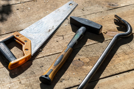 There is a hammer, a crowbar and a saw on a wooden table. The hammer is black, and the crowbar is a large black saw orange. The saw is silver with a black handleの写真素材