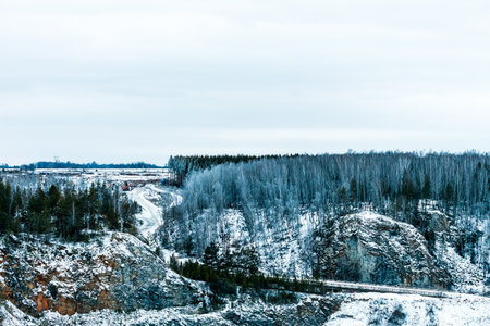 A snowy mountain range with a road running through it. The sky is cloudy and the trees are bare
