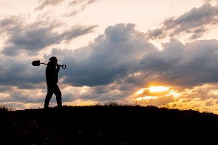 A man is walking on a hill with a shovel in his hand. The sky is cloudy and the sun is settingの写真素材