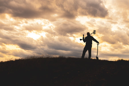 the silhouette of a man against the sunset, a metal detector slung over his shoulder, a shovel in his other hand. in search of metals and coins against the background of sunsetの写真素材