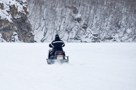 A man is riding a snowmobile on a snowy road. The snowmobile is white and black. The man is wearing a black jacket and a black hat. The snow is falling and the sky is cloudyの写真素材